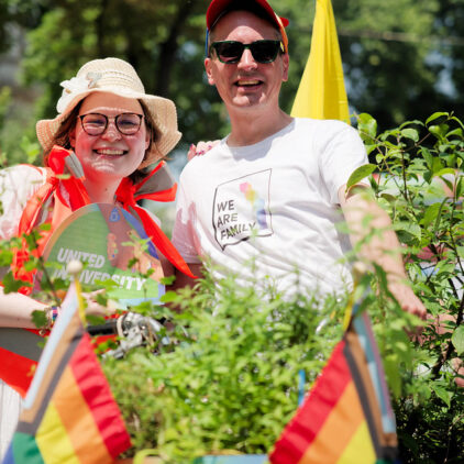 Zwei fröhliche Menschen hinter bepflanztem Rad mit Regenbogenfahnen