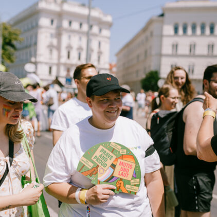 Person (Grüne Andersrum Wien Sprecher:innen Katharina Schöll-Laussermayer) hält „Brave and Proud / Mutig und Laut“ in einer Menschenmenge.