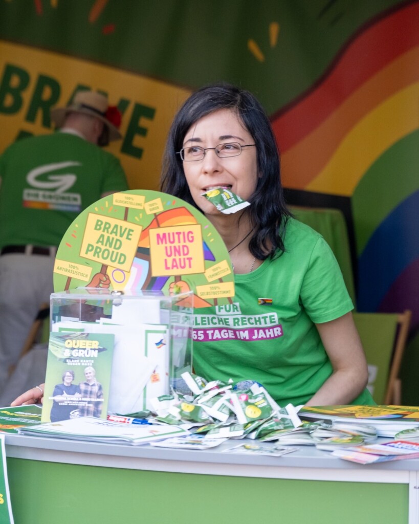 Person am Infostand in grünem Shirt, mit Veganer Gummibärchen und mit Fächer „Brave and Proud / Mutig und laut“ vor Regenbogen-Hintergrund.